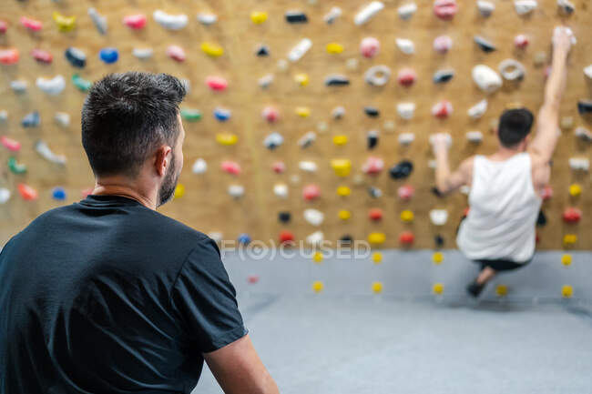 Back view of anonymous male athlete in sportswear watching for climber during workout on wall in gym — Stock Photo