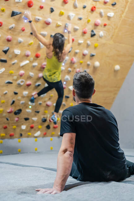 Visão traseira do atleta masculino anônimo em sportswear sentado no chão observando o escalador anônimo borrado durante o treino na parede no ginásio — Fotografia de Stock