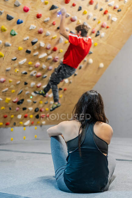 Вид ззаду на невизначену зосереджену жінку в спортивному одязі, що сидить на підлозі і спостерігає за альпіністом на стіні під час тренувань у спортзалі — стокове фото