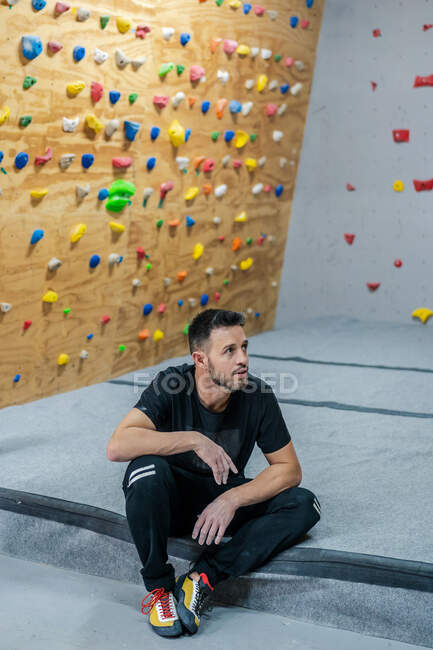 Desde arriba de atleta masculino en ropa deportiva sentado en esteras y mirando hacia otro lado mientras se relaja después de hacer ejercicio de escalada en el gimnasio - foto de stock