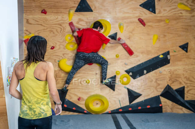 Back view of woman in sportswear standing and watching team male climber on wall in modern gym — Stock Photo