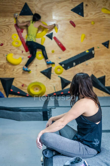 Side view of unrecognizable focused female in sportswear sitting on floor and watching climber on wall during workout in gym — Stock Photo