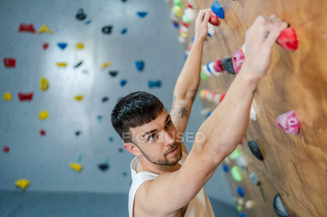 Молодий чоловік у спортивному одязі висить на скелелазінні стінки і дивиться в сторону під час тренувань з боулдингу в спортзалі — стокове фото