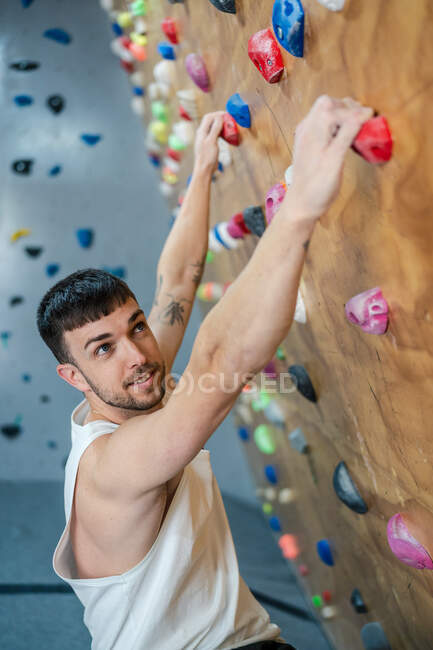 Молодой человек в спортивной одежде висит на стене и смотрит в сторону во время боулдеринга тренировки в тренажерном зале — стоковое фото