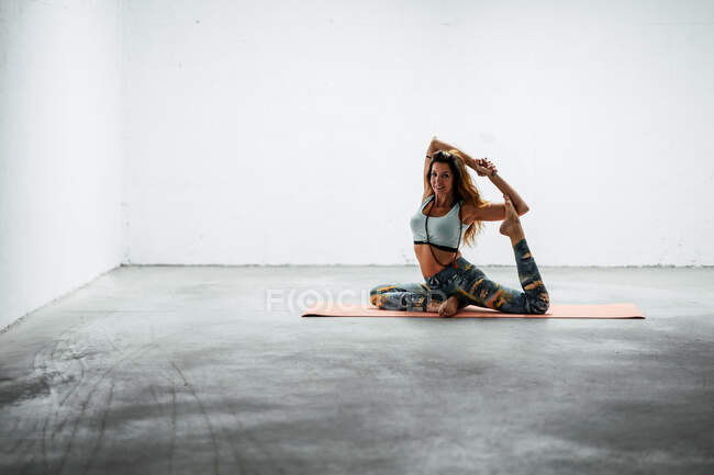 Бічний вид на самицю в спортивному лаві сидить на маті і практикує йогу в Ека Пада Раджакапотасана з жестом мудри — стокове фото
