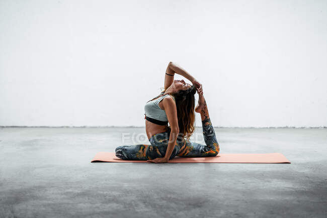 Vista lateral de la hembra en forma de ropa deportiva sentada en la esterilla y practicando yoga en Eka Pada Rajakapotasana con gesto de mudra - foto de stock