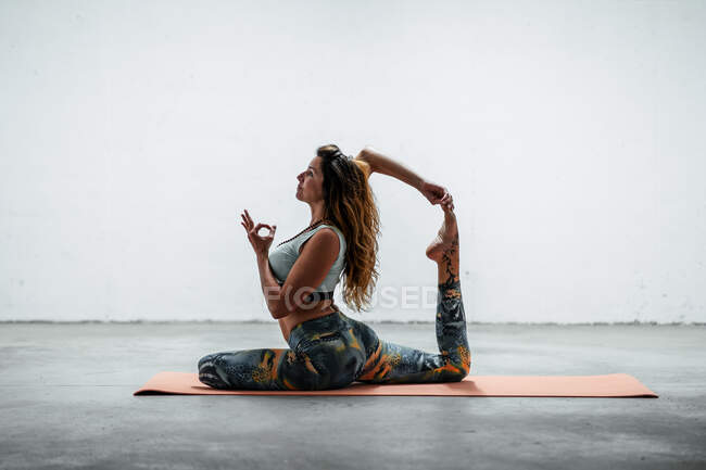 Vista lateral de la hembra en forma de ropa deportiva sentada en la esterilla y practicando yoga en Eka Pada Rajakapotasana con gesto de mudra - foto de stock