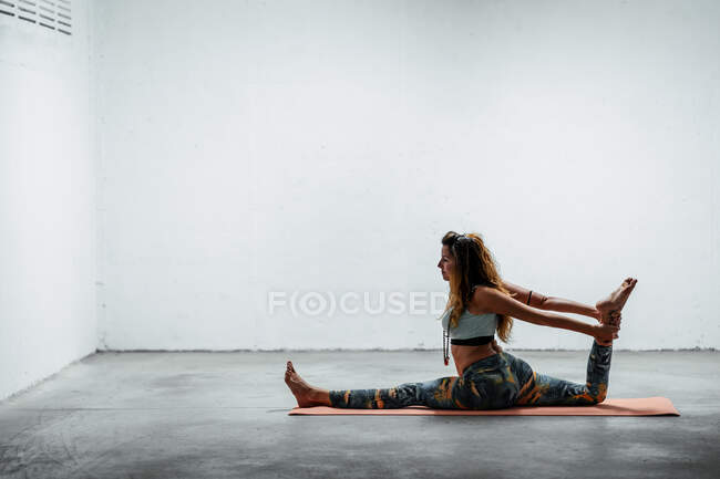 Vista lateral de jovem fêmea flexível em sportswear fazendo frente dividida com perna levantada enquanto sentado no chão — Fotografia de Stock