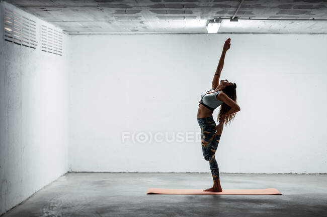 Seitenansicht einer ruhigen Frau mit Sport-Leggings und BH, die auf dem Bein in Urdhva Hastasana balanciert und nach oben schaut — Stockfoto