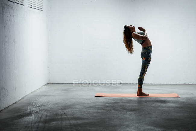 Seitenansicht einer ruhigen Frau mit Sport-Leggings und BH, die auf dem Bein balanciert in stehender Rückenbeuge mit Gebetshänden nach oben blickt — Stockfoto