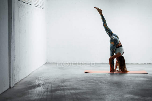 Vista lateral de hembra flexible en desgaste activo de pie sobre estera y haciendo yoga en Urdhva Prasarita Eka Padasana y equilibrio - foto de stock