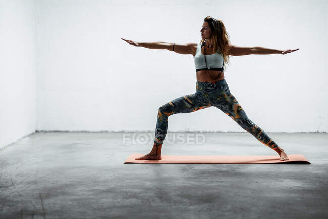 Side view of calm female wearing sports leggings and bra standing on mat warrior two pose looking away — Stock Photo