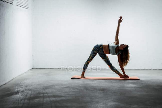 Side view of calm female wearing sports leggings and bra standing on mat in extended triangle pose looking up — Stock Photo