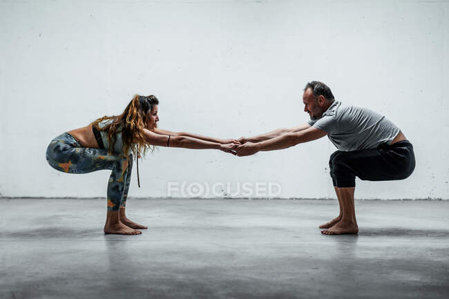 Vista lateral de pareja de enfoque en ropa deportiva practicando yoga en Utkatasana mientras están de pie descalzos en el suelo y mirándose el uno al otro - foto de stock