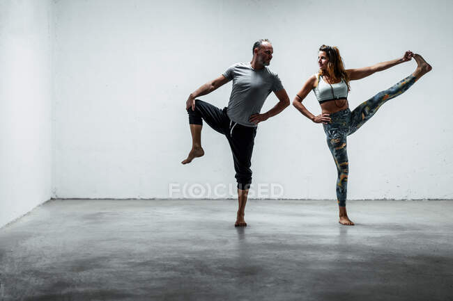 Ein erwachsenes Paar in aktiver Kleidung steht barfuß auf dem Betonboden in Hand to Zeh Asana und schaut sich beim Balancieren auf dem Bein an — Stockfoto