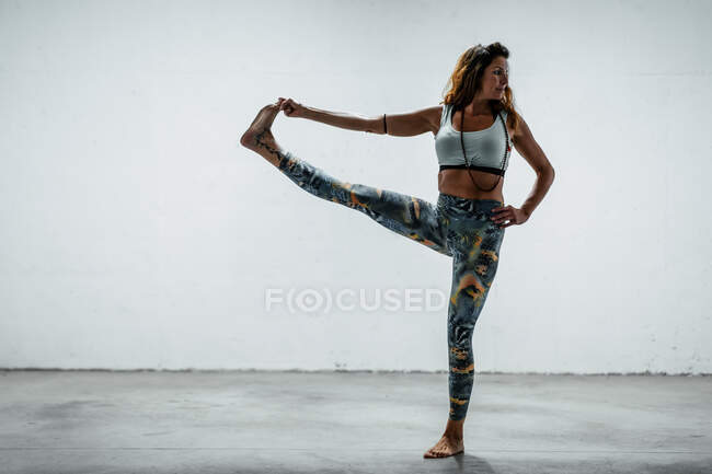 Женщина в активной одежде стоит босиком на бетонном полу в руке к носу и смотрит в сторону, балансируя на ноге — стоковое фото