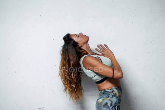 Side view of serene female in sportswear practicing mindfulness and meditating standing with back bend and eyes closed with payer hands on white background — Stock Photo