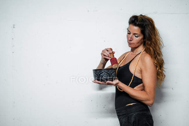Side view serene female wearing rosary standing on white background with Tibetan bowl and playing music for meditation — Stock Photo