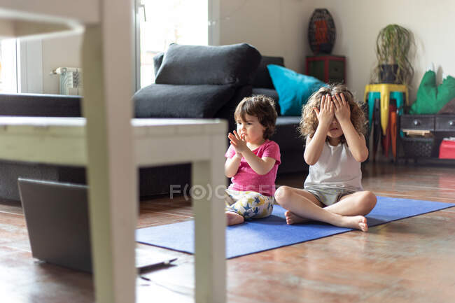 Alto angolo di positive sorelline sedute in posa di loto con le braccia tese mentre guardano video tutorial e praticano yoga insieme in soggiorno a casa — Foto stock