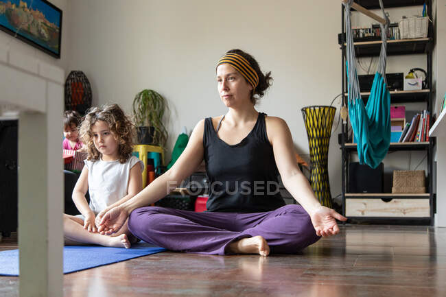 Мать и маленькая дочь в спортивной одежде смотрят онлайн урок йоги и практикуют медитацию в позе лотоса, проводя время вместе дома — стоковое фото