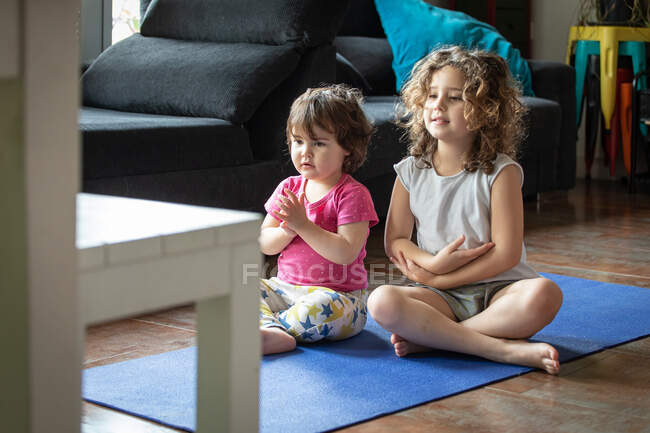 Alto angolo di positive sorelline sedute in posa di loto con le braccia tese mentre guardano video tutorial e praticano yoga insieme in soggiorno a casa — Foto stock