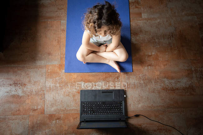 Вид сверху неузнаваемой маленькой девочки, которая смотрит онлайн видео учебник на ноутбуке, сидя на коврике и изучая йогу позировать дома — стоковое фото