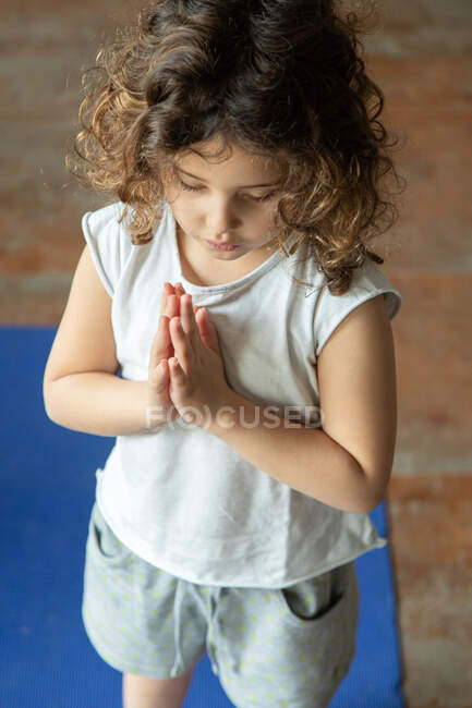 Высокий угол спокойствия маленькая кудрявая девочка в повседневной одежде держа руки в жестах namaste пока стоя на циновке yoga во время урока yoga дома — стоковое фото