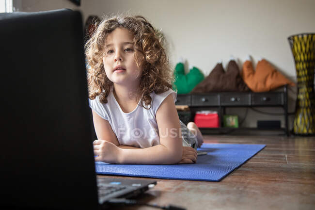 Positive curly haired little girl doing Low Cobra asana while watching video tutorial on laptop at home — Stock Photo