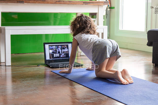Вид ззаду на невпізнавану маленьку дівчинку, яка дивиться онлайн відео-підручник на ноутбуці, сидячи на килимку і вивчаючи йогу позу вдома — стокове фото