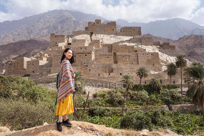 Side view of full body happy ethnic lady in decorative mantle with ornament and yellow dress standing on background of old picturesque Marble Village and rocky mountain ranges — Stock Photo