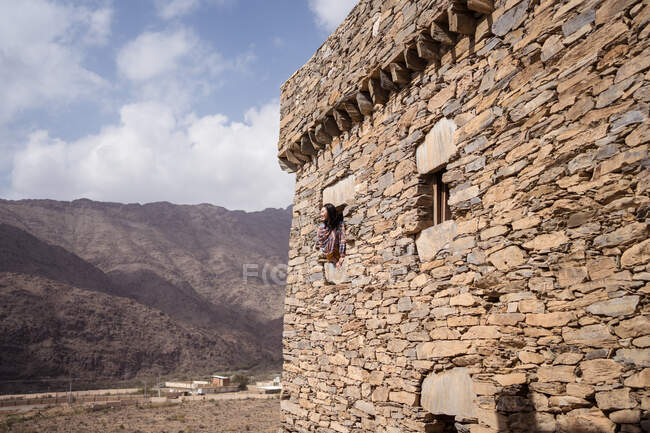 Femme penchée par une fenêtre de l'ancien mur de pierre du bâtiment historique de Marble Village à Al Bahah sur fond de terrain rocheux et ciel nuageux en été en Arabie Saoudite — Photo de stock