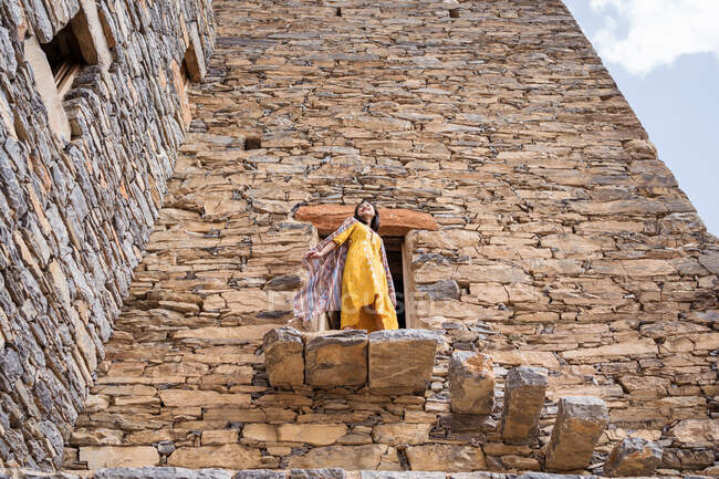 Desde abajo del monumental edificio antiguo con la remota turista femenina que sale de la puerta en vestido amarillo mientras disfruta de un día soleado caliente en Marble Village en Al Bahah - foto de stock
