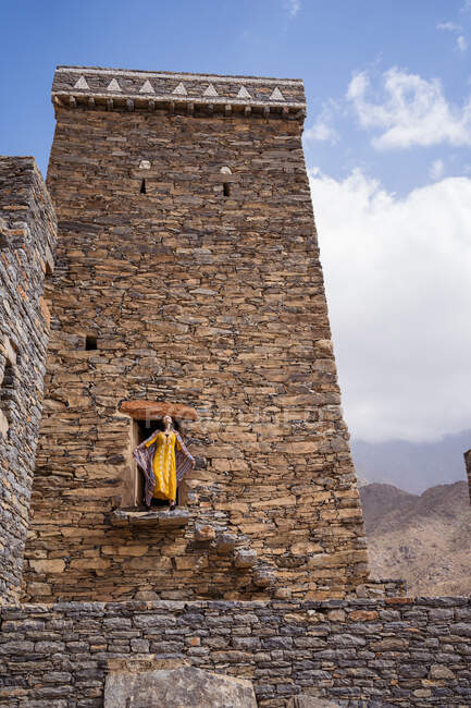 De baixo de monumental edifício antigo com turista feminino remoto saindo da porta em vestido amarelo enquanto desfruta de dia ensolarado quente em Marble Village em Al Bahah — Fotografia de Stock