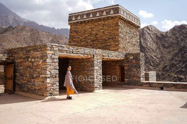Monumental edificio antiguo con el turista femenino remoto que sale de la puerta en vestido amarillo mientras disfruta de un día soleado caliente en Marble Village en Al Bahah - foto de stock