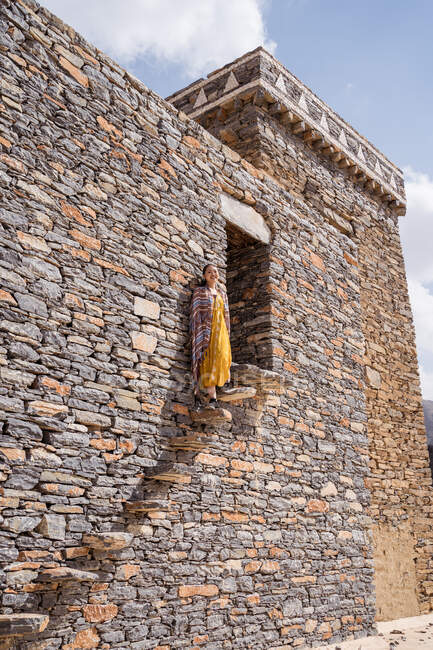 Снизу монументального древнего здания с удаленной туристкой, выходящей из дверей в желтом платье, наслаждаясь жарким солнечным днем в Мраморной деревне в Аль-Баха — стоковое фото