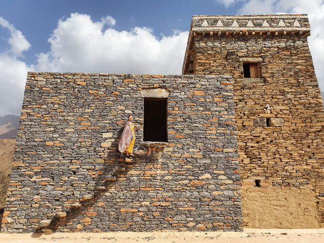 Снизу монументального древнего здания с удаленной туристкой, выходящей из дверей в желтом платье, наслаждаясь жарким солнечным днем в Мраморной деревне в Аль-Баха — стоковое фото