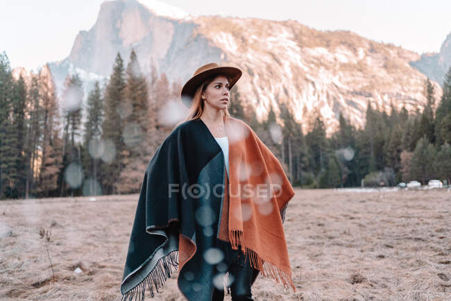 Senderista joven y tranquila en cálido poncho y sombrero de pie en tierra arenosa rodeada de bosque de coníferas y cordillera en el Parque Nacional Yosemite en Estados Unidos - foto de stock