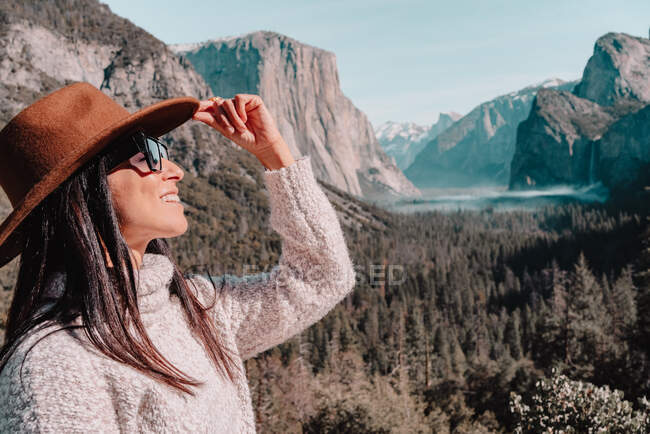 Vista lateral de cuerpo completo de la joven viajera relajada feliz en traje elegante sentada en la frontera de piedra contra pintorescos paisajes de montaña con acantilados rocosos y bosque de coníferas en el Parque Nacional Yosemite en EE.UU. - foto de stock