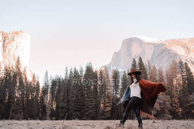 Vue latérale de la jeune femme gaie dans le poncho et chapeau marchant sur l'herbe sèche près de la forêt avec des falaises rocheuses de granit en arrière-plan dans la journée ensoleillée dans le parc national Yosemite aux États-Unis — Photo de stock