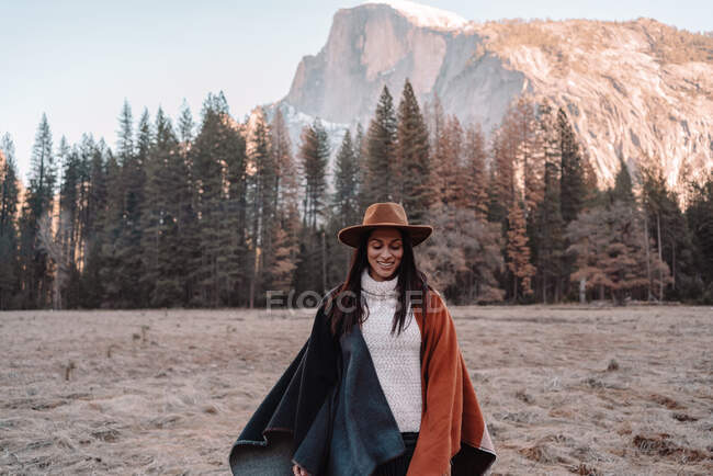 Feliz relaxado jovem viajante feminino em roupa elegante sentado na fronteira de pedra contra a paisagem pitoresca montanha com falésias rochosas e floresta de coníferas no Parque Nacional de Yosemite, nos EUA — Fotografia de Stock