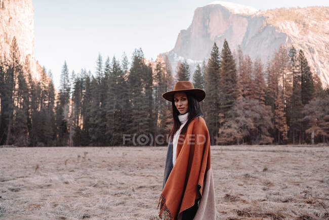 Feliz viajero joven relajado en traje elegante sentado en la frontera de piedra contra pintorescos paisajes de montaña con acantilados rocosos y bosque de coníferas en el Parque Nacional Yosemite en Estados Unidos - foto de stock