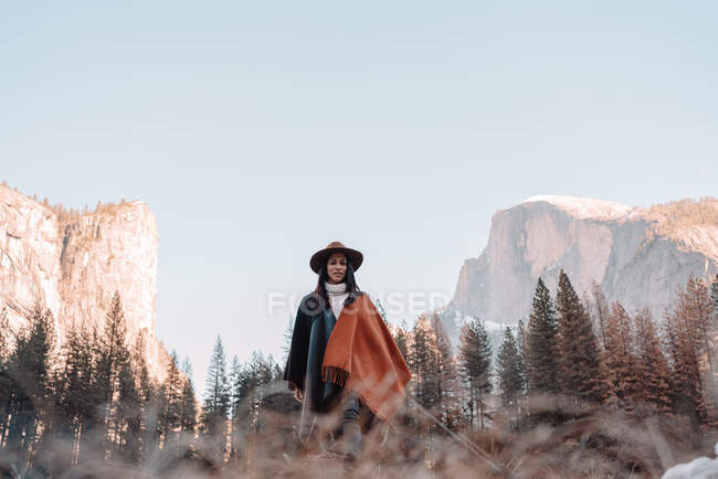 Felice rilassato giovane viaggiatore femminile in abito elegante seduto sul confine di pietra contro pittoresco scenario di montagna con scogliere rocciose e foresta di conifere nel Parco Nazionale di Yosemite negli Stati Uniti — Foto stock