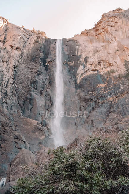 Снизу удивительный вид на мощный водопад, струящийся с высокой скалистой скалы против безоблачного неба в солнечный день в Национальном парке Йосемити в США — стоковое фото