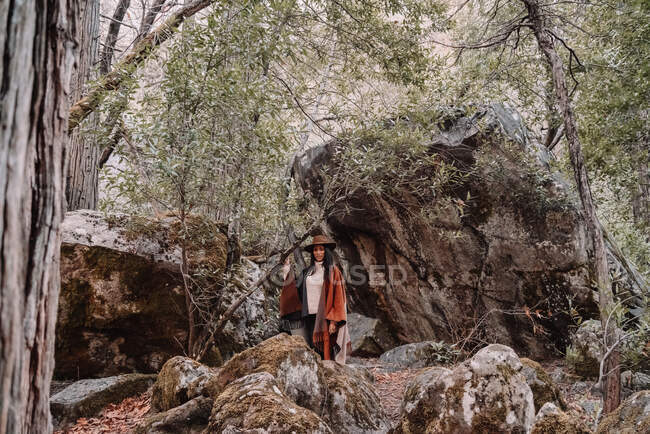 Junge Wanderin in stylischer Boho-Kleidung und Hut steht zwischen riesigen Felsbrocken im Wald während einer Reise zum Yosemite-Nationalpark in Kalifornien — Stockfoto