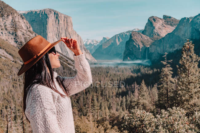 Vista laterale del corpo pieno di felice rilassato giovane viaggiatore femminile in abito elegante seduto sul confine di pietra contro pittoreschi paesaggi di montagna con scogliere rocciose e foresta di conifere nel Parco Nazionale di Yosemite negli Stati Uniti — Foto stock