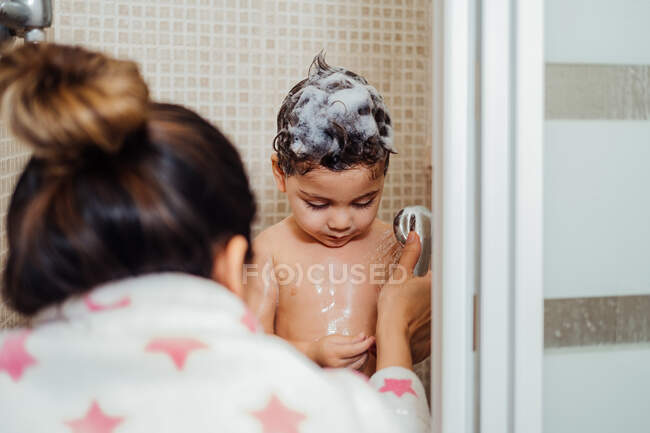 Visão traseira da mulher de roupão de banho em pé no banheiro e lavar a criança com espuma no cabelo — Fotografia de Stock
