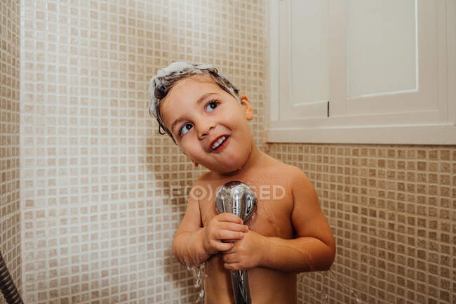 Bambino sorridente con schiuma sulla testa in piedi in bagno con doccia e canto mentre distoglie lo sguardo — Foto stock