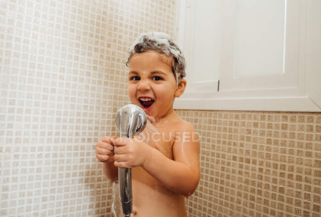Niño sonriente con espuma en la cabeza de pie en el baño con ducha y cantando mientras mira la cámara - foto de stock