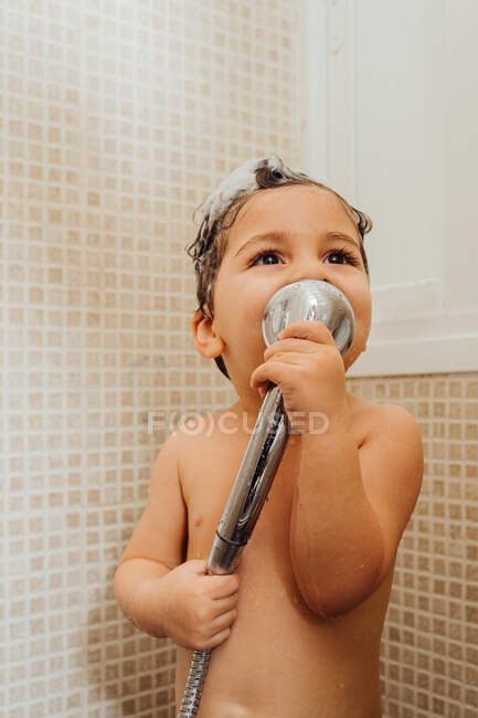 Улыбающийся маленький ребенок с пеной на голове стоит в ванной комнате с душем и поет, отводя взгляд — стоковое фото