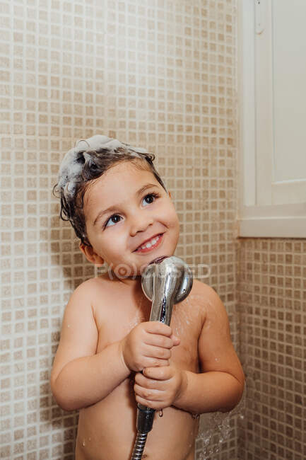 Criança sorridente com espuma na cabeça em pé no banheiro com chuveiro e cantando enquanto olha para longe — Fotografia de Stock
